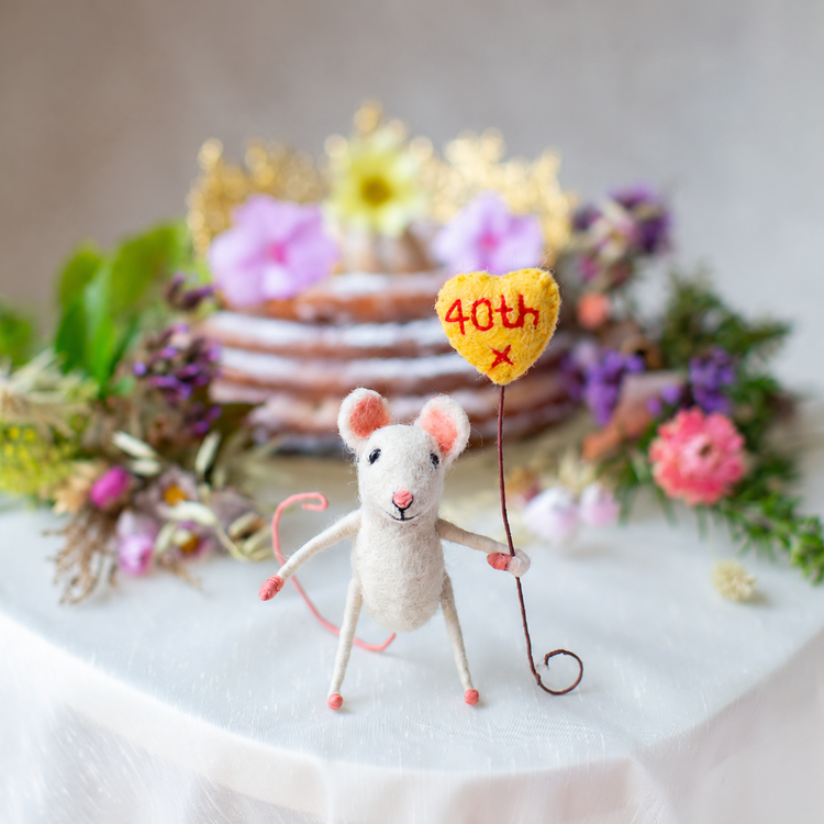 A small felt mouse figurine stands on a white tablecloth holding a yellow heart-shaped balloon that reads &