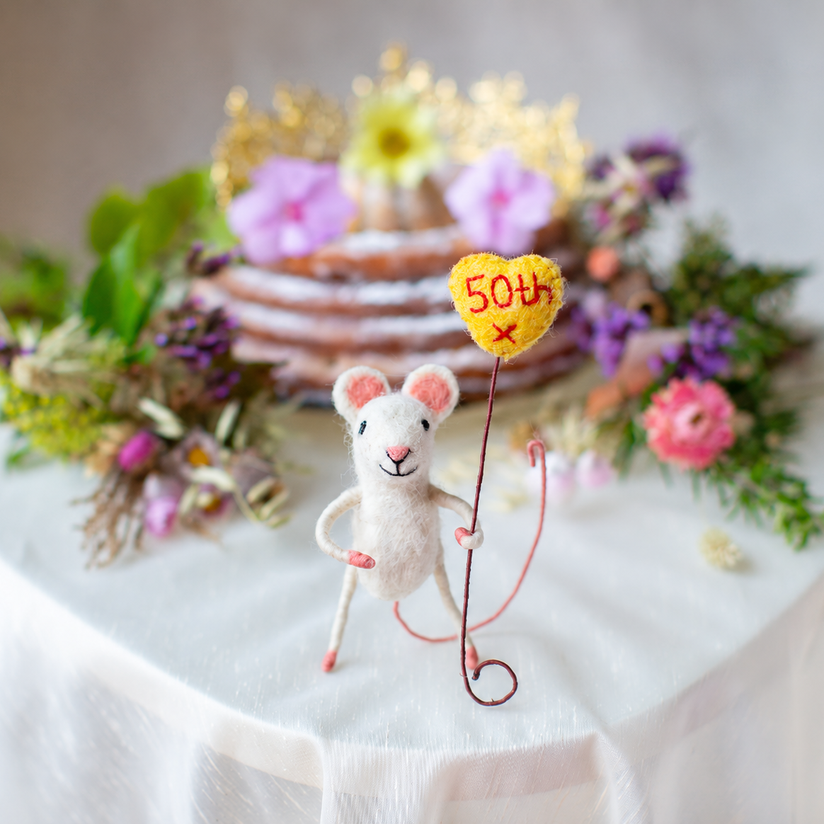A small white felt mouse figurine holding a yellow heart-shaped balloon that says “50th” stands on a white draped table. In the softly blurred background is a decorated cake with pink and yellow flowers.